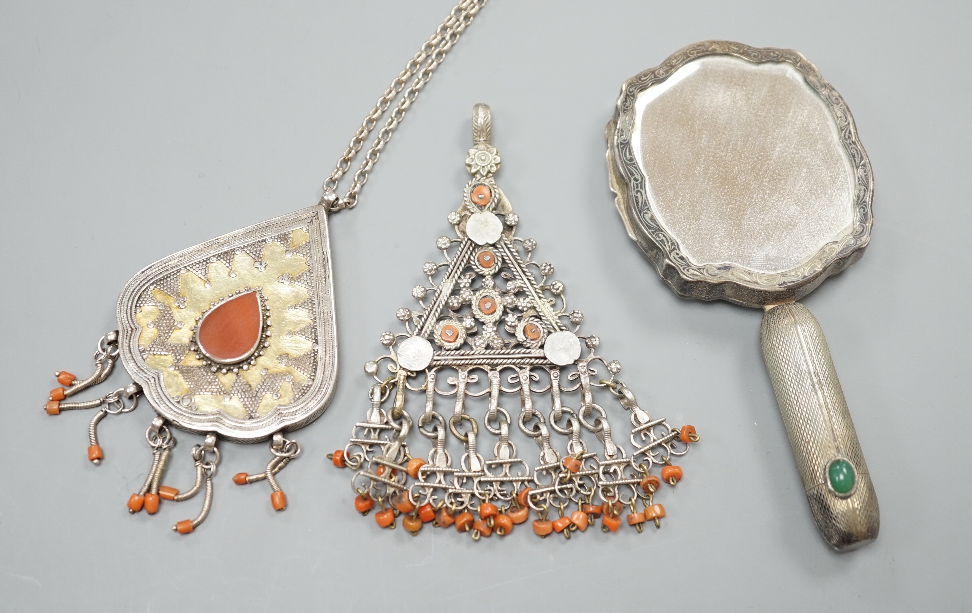 Two shaped Afghan? white metal or parcel gilt white metal, coral bead set pendants, one on a white metal chain, together with a continental engine turned white metal minaudiere, modelled as a hand mirror, 12.3cm